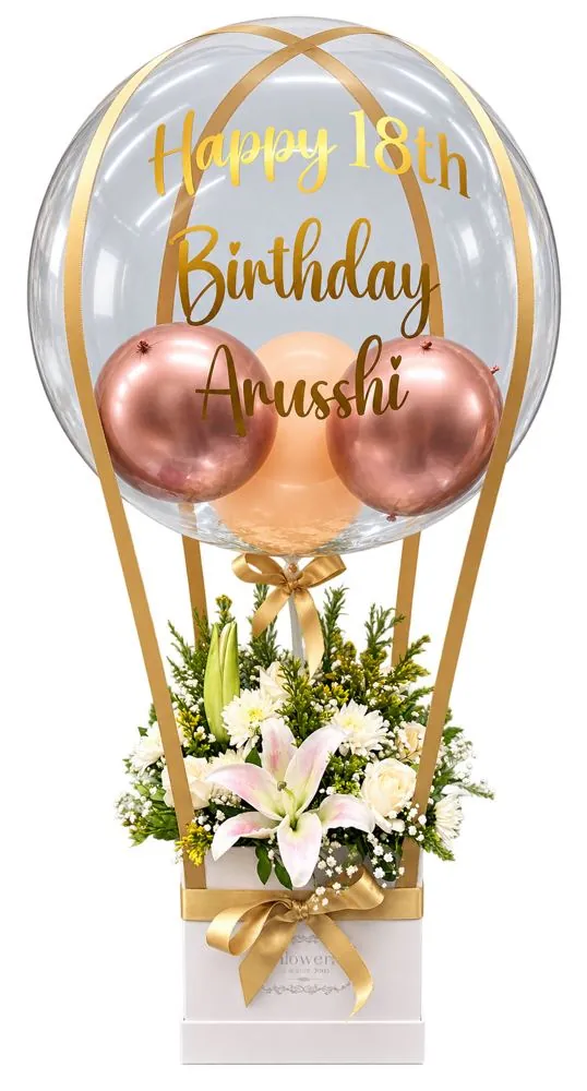 Bubble balloon with rose gold balloons inside, gold Happy 18th Birthday lettering and gold ribbons forming a hot air balloon over a white flower box